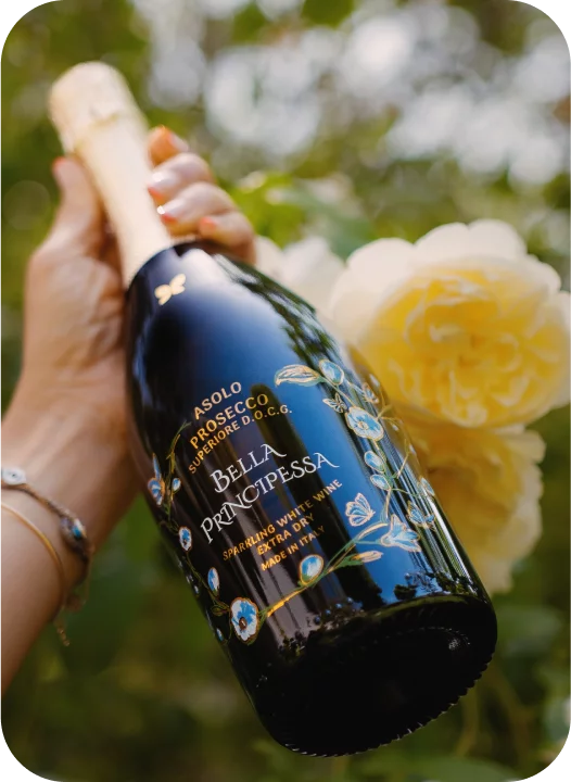 Gepflegte Hand hält eine Flasche Luxus-Prosecco Bella Principessa vor blühenden Rosen – Warum ist Prosecco so günstig?